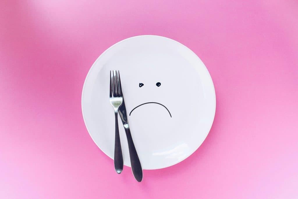 Des couverts sont posées sur une assiette sur fond rose. Un visage triste est dessiné sur l'assiette. Ceci représente le dégoût que peuvent provoquer certains aliments.