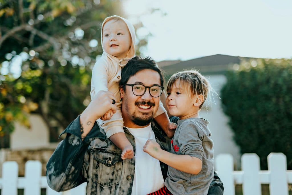 Un homme avec des lunettes porte un enfant dans ses bras et un enfant plus jeune sur ses épaules